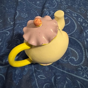 May include: A yellow ceramic teapot with a matching handle and spout. The lid is light purple with a ruffled edge and a small, round, orange knob. The teapot is set against a blue patterned background.
