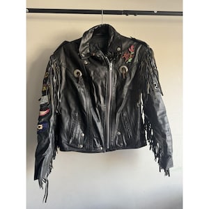 May include: Black leather biker jacket with fringing on the sleeves and back. The jacket has silver-tone metal hardware, including zips and press studs, and embroidered patches. A rose and the words "Lady Luck" are embroidered on the upper chest.