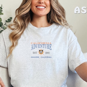 May include: A gray t-shirt with a graphic print of the California Adventure logo. The logo features a sun with Mickey Mouse's head in the center. The text reads "California Adventure Est. 2001 Anaheim, California".