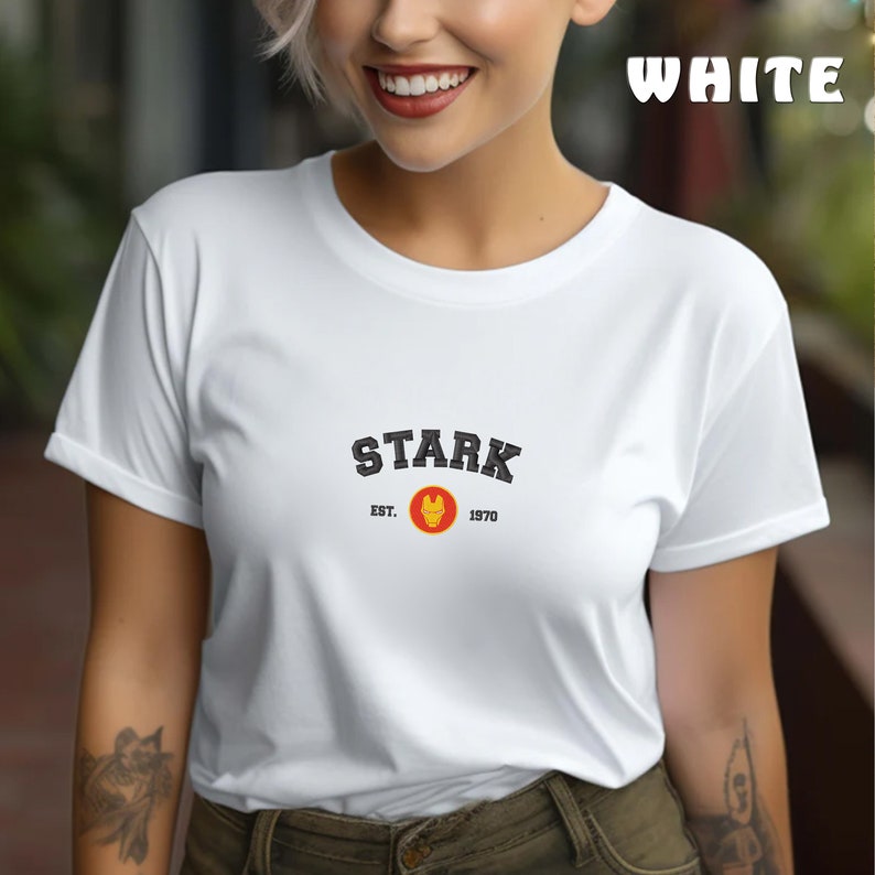 以下が含まれることがあります： 白いTシャツに、黒い文字で「STARK」と書いてあり、その下に「EST. 1970」と書いてあります。グラフィックには、赤と黄色のアイアンマンのロゴも含まれています。