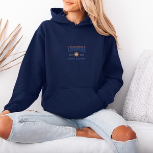 Puede incluir: Sudadera con capucha azul marino con bolsillo delantero y capucha con cord&oacute;n. La sudadera presenta el texto "CALIFORNIA ADVENTURE" con un gr&aacute;fico de sol y el texto "ANAHEIM, CALIFORNIA". La sudadera est&aacute; hecha de un material suave.