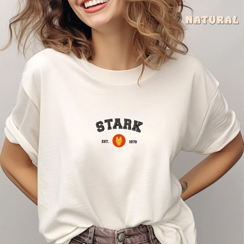 以下が含まれることがあります： 白いTシャツに、黒い文字で「STARK EST. 1970」と、小さく黄色と赤のアイアンマンのロゴが書かれている。
