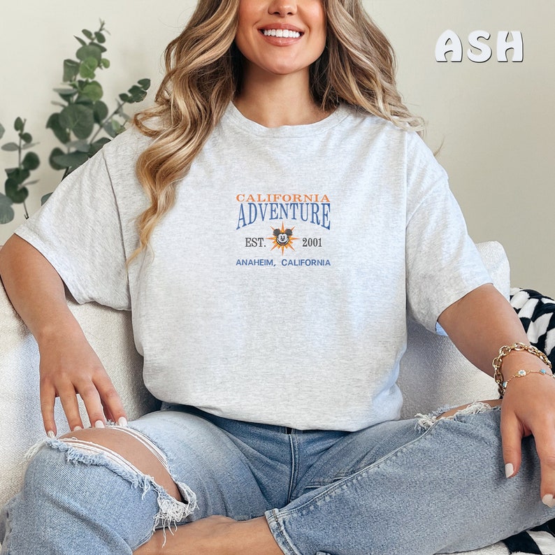 May include: A gray t-shirt with a graphic print of the California Adventure logo. The logo features a sun with Mickey Mouse's head in the center. The text reads "California Adventure Est. 2001 Anaheim, California".