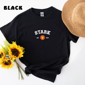 以下が含まれることがあります： 黒のTシャツに、白い「STARK」のグラフィックと、赤と黄色のアイアンマンのロゴがプリントされています。グラフィックの下のテキストは「EST. 1970」です。
