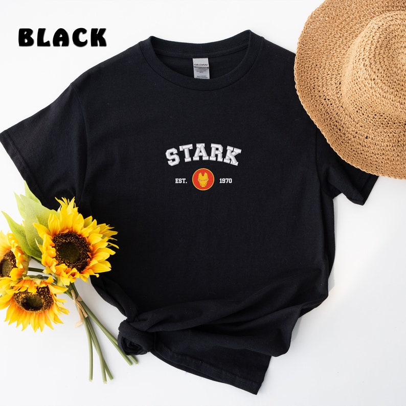 以下が含まれることがあります： 黒のTシャツに、白い「STARK」のグラフィックと、赤と黄色のアイアンマンのロゴがプリントされています。グラフィックの下のテキストは「EST. 1970」です。