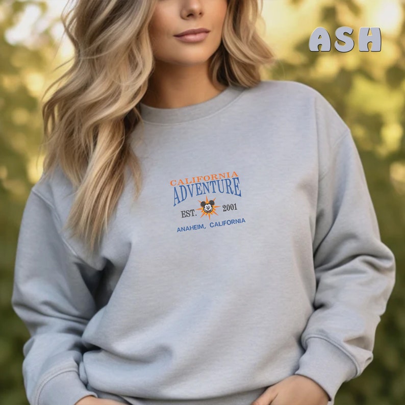 May include: A light gray sweatshirt with a blue and orange graphic print that says "California Adventure Est. 2001 Anaheim, California" with a Mickey Mouse head inside a sun graphic.