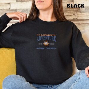 May include: Black sweatshirt with a graphic print of a yellow sun with a Mickey Mouse head in the center. The text "California Adventure" is above the sun, and the text "Est. 2001 Anaheim, California" is below the sun.