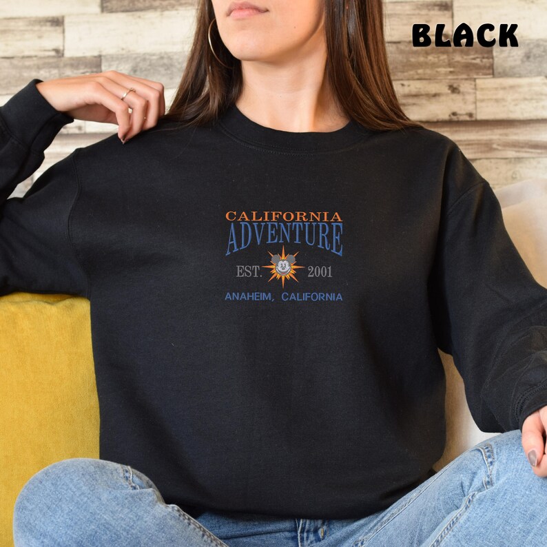 May include: Black sweatshirt with a graphic print of a yellow sun with a Mickey Mouse head in the center. The text "California Adventure" is above the sun, and the text "Est. 2001 Anaheim, California" is below the sun.