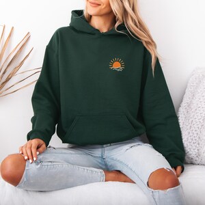 May include: A dark green hoodie with a front pocket and a hood. The hoodie features a small embroidered design of a sun setting over waves on the upper left chest. The person is wearing light blue jeans with ripped knees.