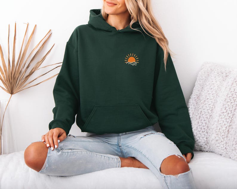 May include: A dark green hoodie with a front pocket and a hood. The hoodie features a small embroidered design of a sun setting over waves on the upper left chest. The person is wearing light blue jeans with ripped knees.