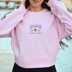May include: A light pink sweatshirt with the text "CALIFORNIA ADVENTURE EST. 2001 ANAHEIM, CALIFORNIA" and a cartoon sun with Mickey Mouse's head in the center.