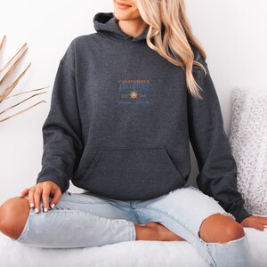Puede incluir: Sudadera con capucha gris carb&oacute;n con bolsillo delantero y capucha. La sudadera tiene un dise&ntilde;o en el pecho que dice "California Adventure" con una rosa de los vientos y un gr&aacute;fico de sol. La persona lleva vaqueros azul claro.