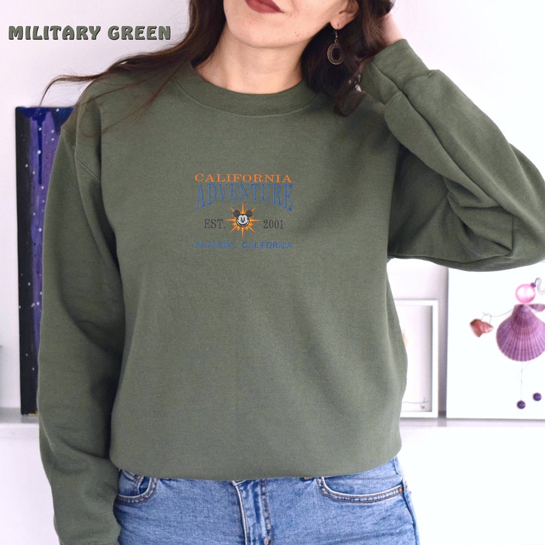 May include: A green sweatshirt with the text "CALIFORNIA ADVENTURE EST. 2001 ANAHEIM, CALIFORNIA" and a cartoon sun with a Mickey Mouse face.