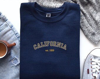 Peut inclure: Un sweat-shirt bleu marine avec un graphisme brodé en or qui dit "CALIFORNIA est. 1850".