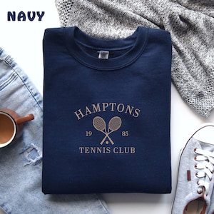 May include: Navy blue sweatshirt with a gold graphic of two tennis rackets crossed with a tennis ball in the center. The text "HAMPTONS 19 85 TENNIS CLUB" is printed below the rackets.