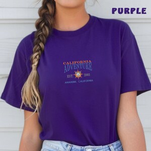 May include: Purple t-shirt with a graphic of a sun with the text "California Adventure Est. 2001 Anaheim, California" printed on the front.