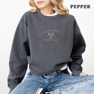 May include: A charcoal gray sweatshirt with a vintage-inspired design featuring the text "HAMPTONS TENNIS CLUB" and a pair of crossed tennis rackets.