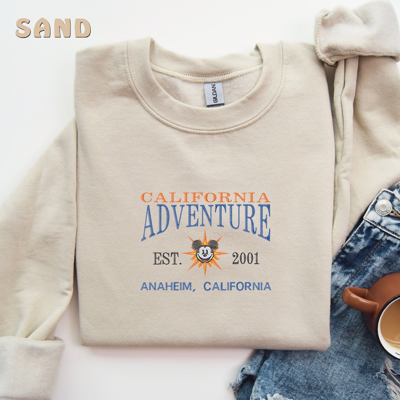 May include: A beige sweatshirt with a vintage-style graphic print that says "California Adventure Est. 2001 Anaheim, California" with a cartoon Mickey Mouse head in the center.
