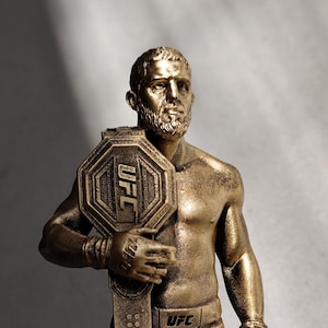 Puede incluir: Una figura dorada de un luchador barbudo sin camisa, con pantalones cortos deportivos y vendas en las manos. La figura sostiene un gran cinturón de campeonato de la UFC octogonal sobre el pecho. El logo de la UFC aparece en el cinturón y el pantalón.