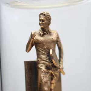 May include: A gold-colored statue of a tennis player in a victory pose, holding a racket. The figure is wearing a collared shirt and shorts. The statue is inside a clear glass container.