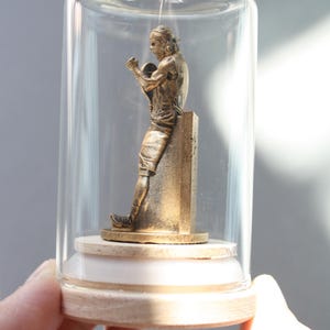 May include: A bronze-colored statue of a boxer inside a glass cloche. The figure is posed mid-fight, leaning against a pillar. The cloche sits on a wooden base, held by a hand.