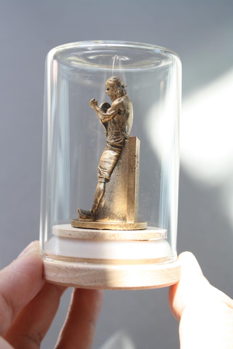 May include: A bronze-colored statue of a boxer inside a glass cloche. The figure is posed mid-fight, leaning against a pillar. The cloche sits on a wooden base, held by a hand.