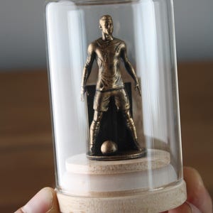 May include: A bronze-colored soccer player figurine inside a glass cloche. The figurine is standing with a soccer ball at its feet. The cloche has a wooden base and a clear glass dome.