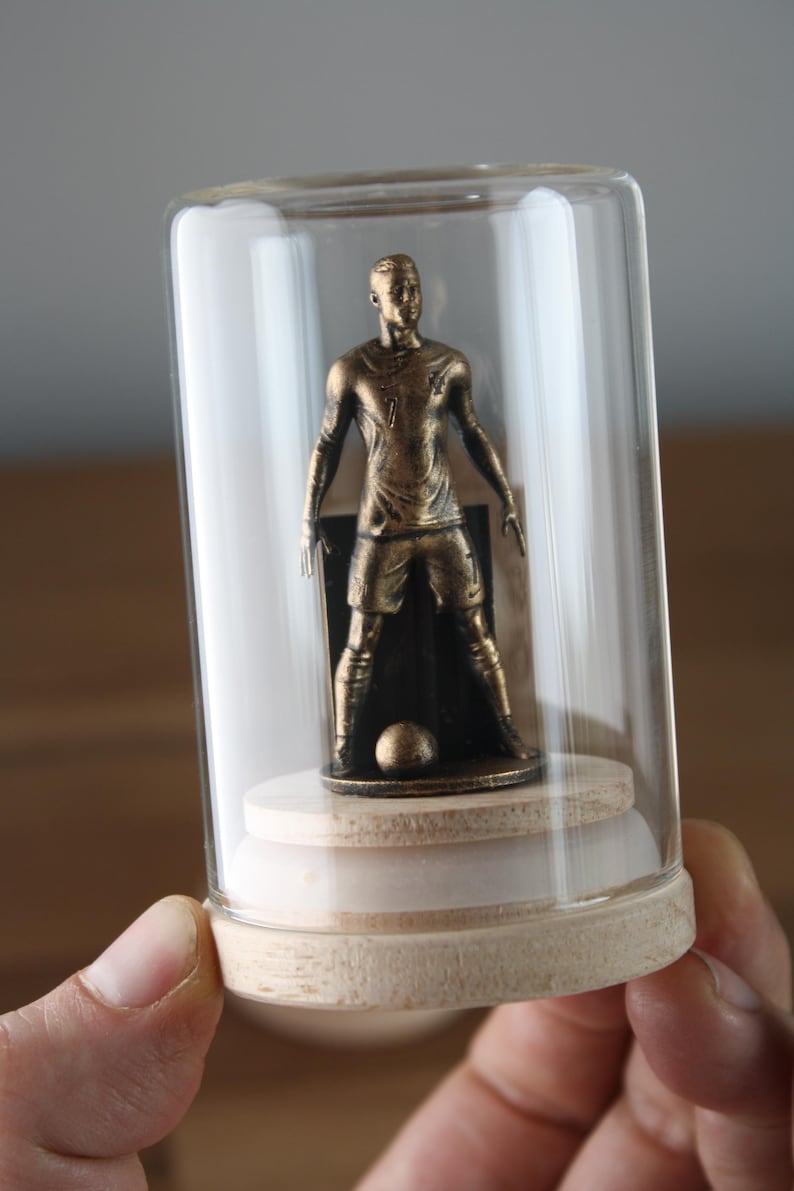 May include: A bronze-colored soccer player figurine inside a glass cloche. The figurine is standing with a soccer ball at its feet. The cloche has a wooden base and a clear glass dome.