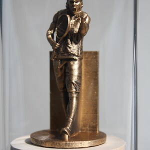 May include: A bronze-colored tennis player figurine inside a glass dome. The figurine is posed mid-swing, holding a racket, and is mounted on a round base. The figure is approximately 5 inches tall.