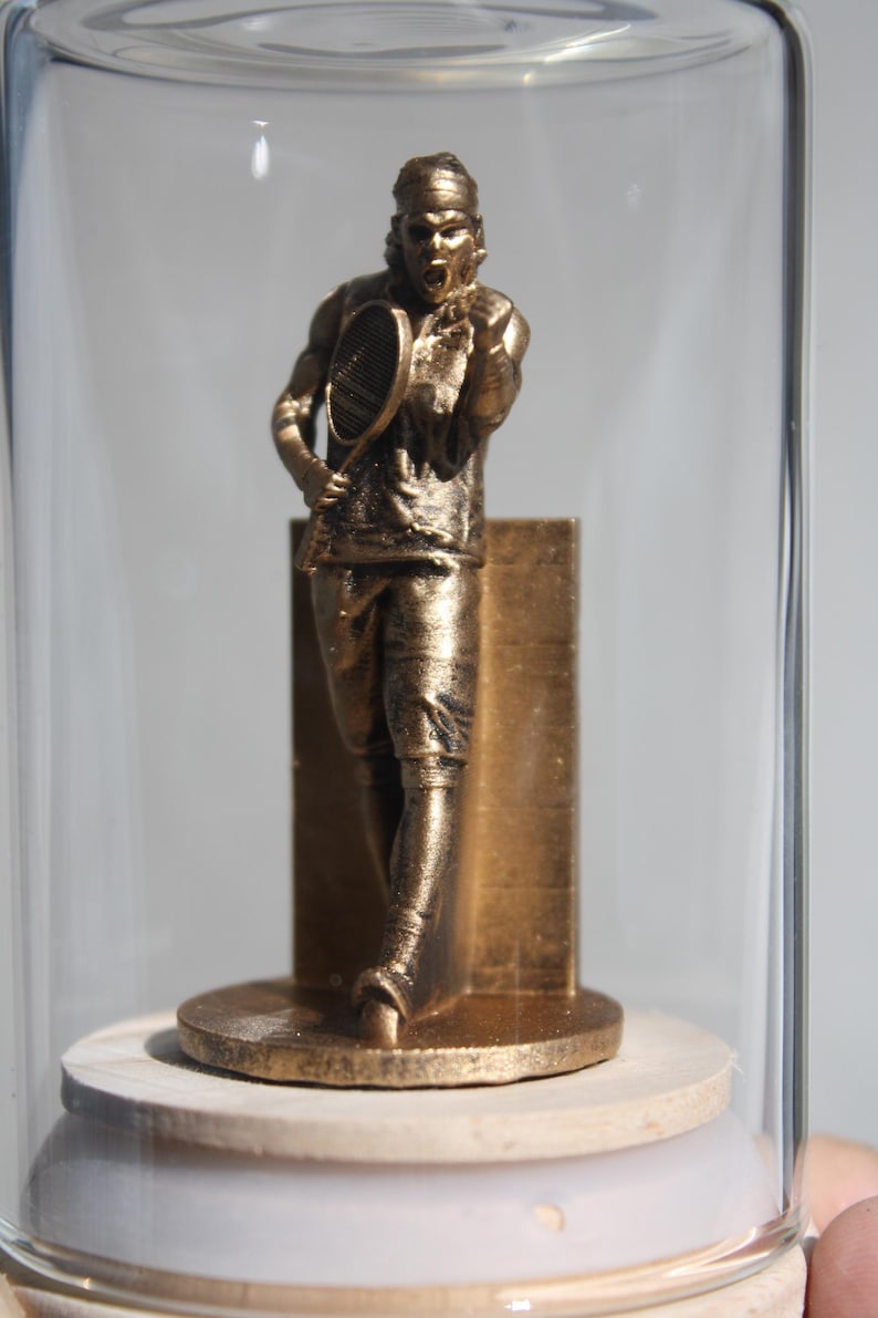 May include: A bronze-colored tennis player figurine inside a glass dome. The figurine is posed mid-swing, holding a racket, and is mounted on a round base. The figure is approximately 5 inches tall.