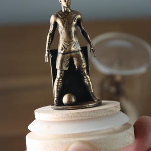 May include: A bronze-colored soccer player figurine on a wooden base. The figure is posed with arms at the sides, wearing a jersey with the number 7. A soccer ball is at the player's feet. The base has a layered design.