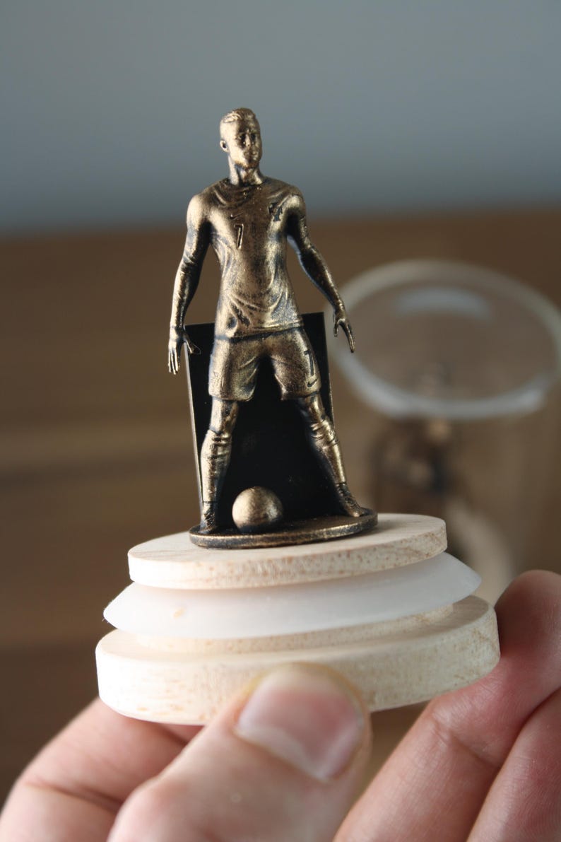 May include: A bronze-colored soccer player figurine on a wooden base. The figure is posed with arms at the sides, wearing a jersey with the number 7. A soccer ball is at the player's feet. The base has a layered design.