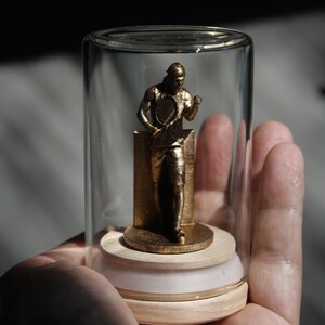 May include: A gold-colored tennis player figurine inside a clear glass jar. The figurine is standing on a small, round base. The jar is held in a person's hand.