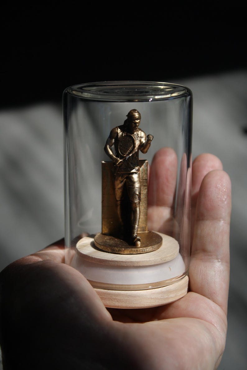 May include: A gold-colored tennis player figurine inside a clear glass jar. The figurine is standing on a small, round base. The jar is held in a person's hand.