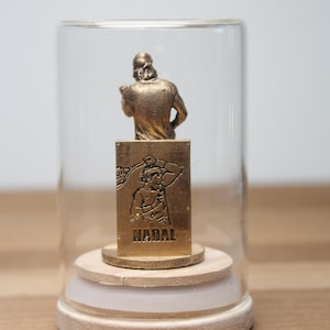 May include: A gold-colored figurine of a person in a glass dome. The figurine is mounted on a wooden base. The base has a gold-colored plaque with the name "NADAL" and an image of a person. The dome is clear.