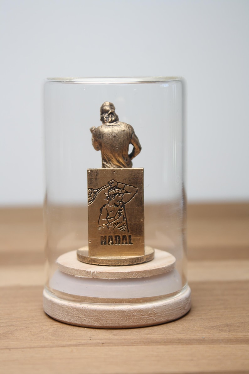 May include: A gold-colored figurine of a person in a glass dome. The figurine is mounted on a wooden base. The base has a gold-colored plaque with the name "NADAL" and an image of a person. The dome is clear.
