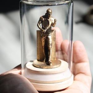 May include: A gold-colored tennis player figurine inside a clear glass display case. The figurine is standing on a wooden base with a white rim. The tennis player is holding a tennis racket.