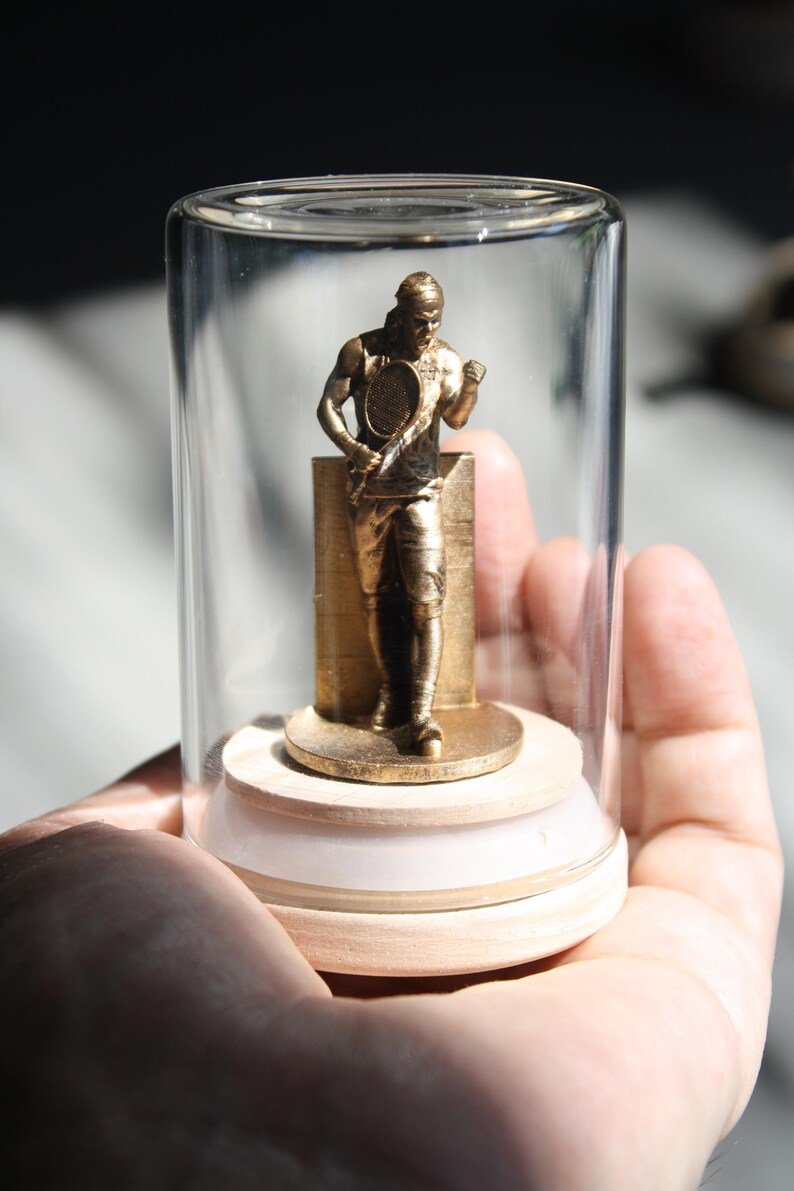 May include: A gold-colored tennis player figurine inside a clear glass display case. The figurine is standing on a wooden base with a white rim. The tennis player is holding a tennis racket.