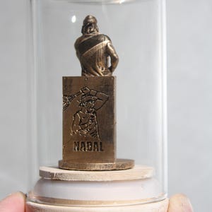 May include: A bronze-colored figurine of a tennis player inside a glass display case. The figurine is mounted on a wooden base. The word "NADAL" is engraved on the front of the base.