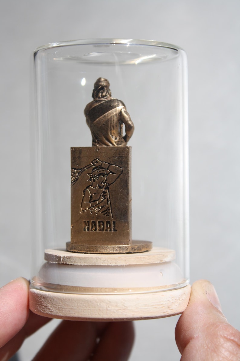 May include: A bronze-colored figurine of a tennis player inside a glass display case. The figurine is mounted on a wooden base. The word "NADAL" is engraved on the front of the base.