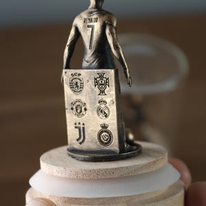 May include: A bronze-colored figurine of a soccer player wearing the number 7 and the name "Ronaldo" on the back. The figurine holds a plaque with team logos. The figurine is mounted on a wooden base with a white rubber ring.