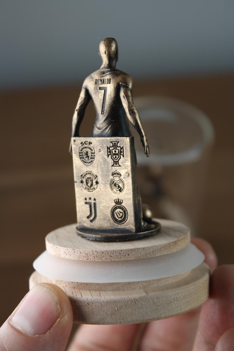 May include: A bronze-colored figurine of a soccer player wearing the number 7 and the name "Ronaldo" on the back. The figurine holds a plaque with team logos. The figurine is mounted on a wooden base with a white rubber ring.