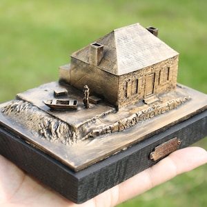May include: A bronze model of a small house with a boat in front of it. The model is on a black wooden base with a plaque that reads "Historic Site 1768".