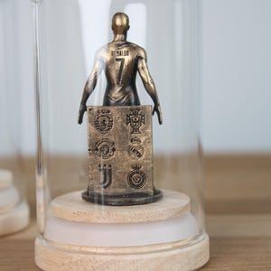 May include: A bronze-colored figurine of a soccer player wearing the number 7 and the name "Ronaldo" on the back, displayed under a glass cloche. The figurine is mounted on a wooden base with a white accent and features team logos.