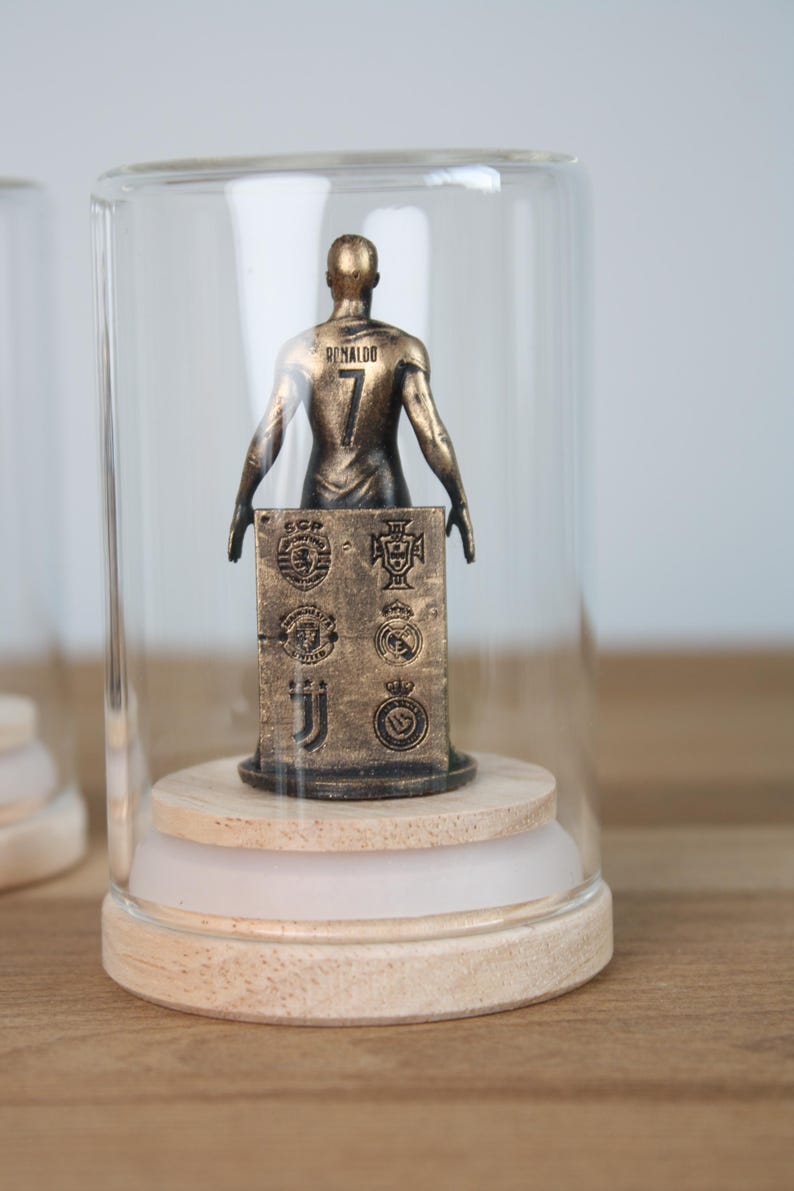 May include: A bronze-colored figurine of a soccer player wearing the number 7 and the name "Ronaldo" on the back, displayed under a glass cloche. The figurine is mounted on a wooden base with a white accent and features team logos.