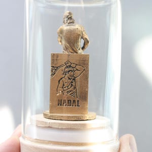 May include: A miniature gold-colored statue of a tennis player inside a glass dome. The statue is mounted on a wooden base. The base has the word "NADAL" engraved on it. The dome is held by a person's hand.