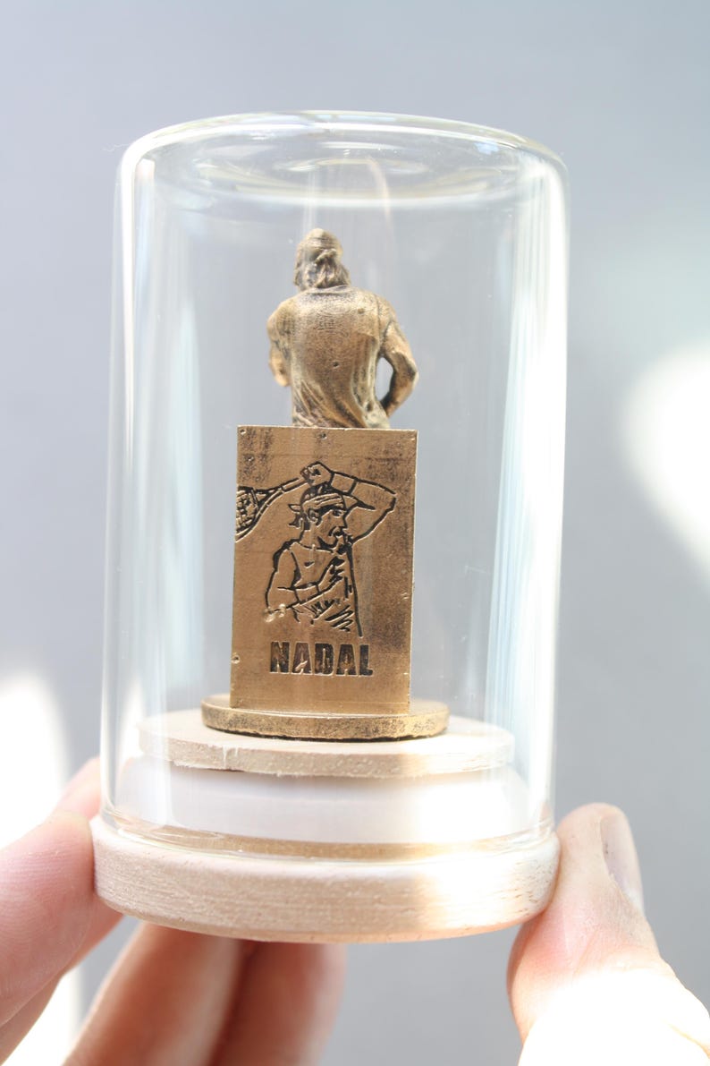May include: A miniature gold-colored statue of a tennis player inside a glass dome. The statue is mounted on a wooden base. The base has the word "NADAL" engraved on it. The dome is held by a person's hand.