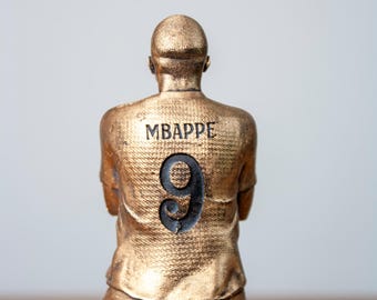May include: A gold-coloured figurine of a football player wearing a jersey with the name "MBAPPE" and the number "9" on the back.