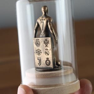 May include: A bronze-colored figurine of a soccer player inside a glass dome. The player wears a jersey with the number 7 and the name "Ronaldo" on the back. A small plaque displays team logos. The figurine sits on a wooden base.