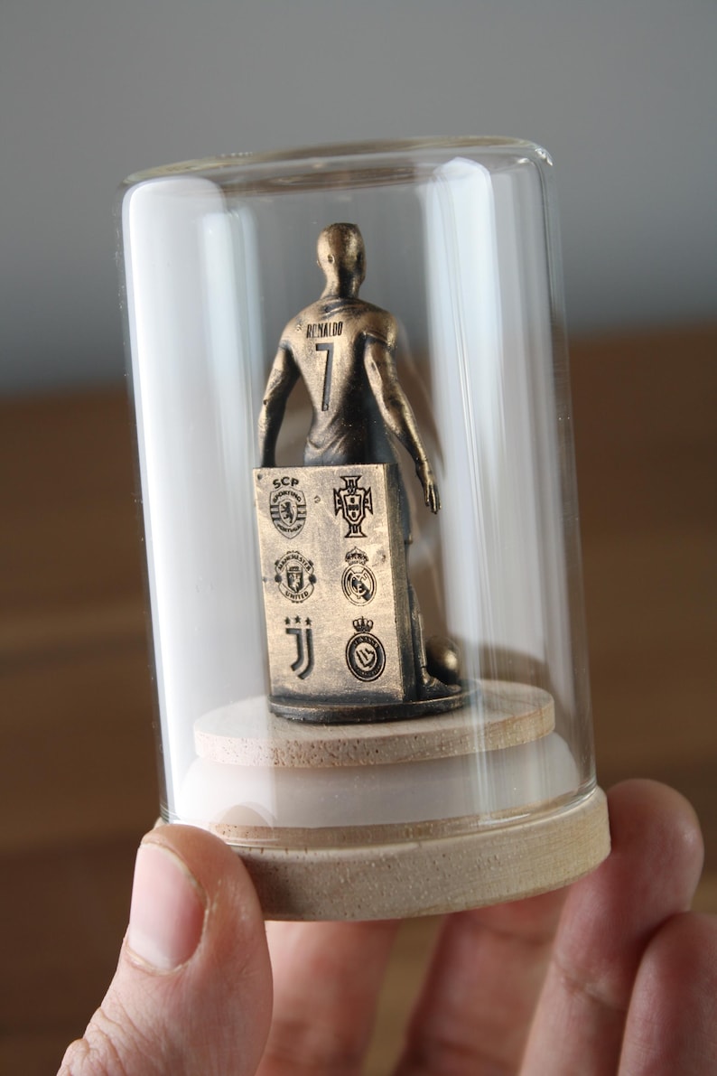May include: A bronze-colored figurine of a soccer player inside a glass dome. The player wears a jersey with the number 7 and the name "Ronaldo" on the back. A small plaque displays team logos. The figurine sits on a wooden base.